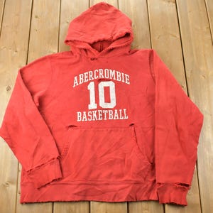 May include: Red hooded sweatshirt with distressed detailing. The front features white text that reads "Abercrombie 10 Basketball".