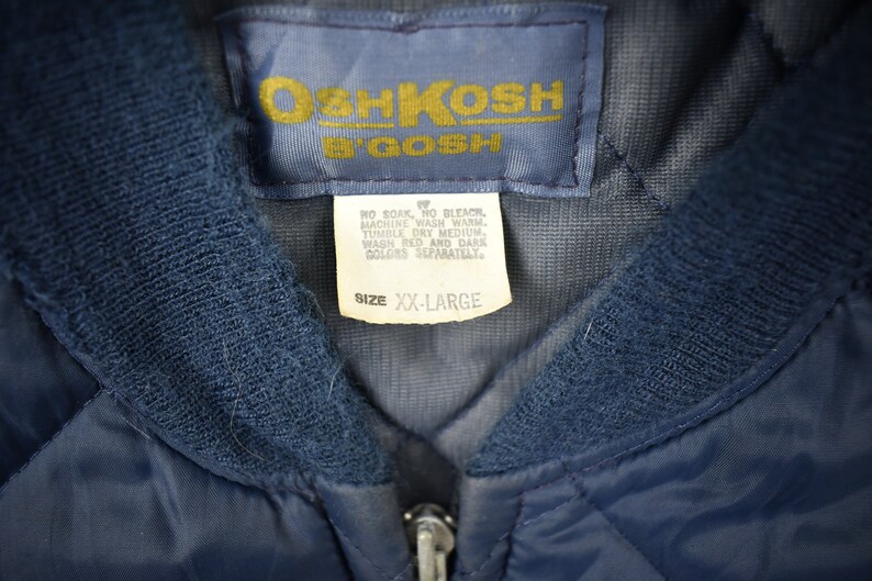 May include: A navy blue quilted jacket with a zipper closure and a label that reads "OshKosh B'Gosh" and "Size XX-Large". The label also includes care instructions.