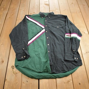 Puede incluir: Una camisa de manga larga con botones con un diseño de bloques de color negro y verde. La camisa tiene un patrón de rayas blancas y moradas en el lado izquierdo y un patrón de rayas blancas y verdes en la manga derecha.