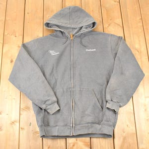 Puede incluir: Una sudadera gris con cremallera, capucha y bolsillo delantero. El logotipo de Carhartt es visible en el pecho derecho. La sudadera tiene un cord&oacute;n y pu&ntilde;os y dobladillo acanalados. El texto "Power Equipment Company" est&aacute; en el pecho izquierdo.