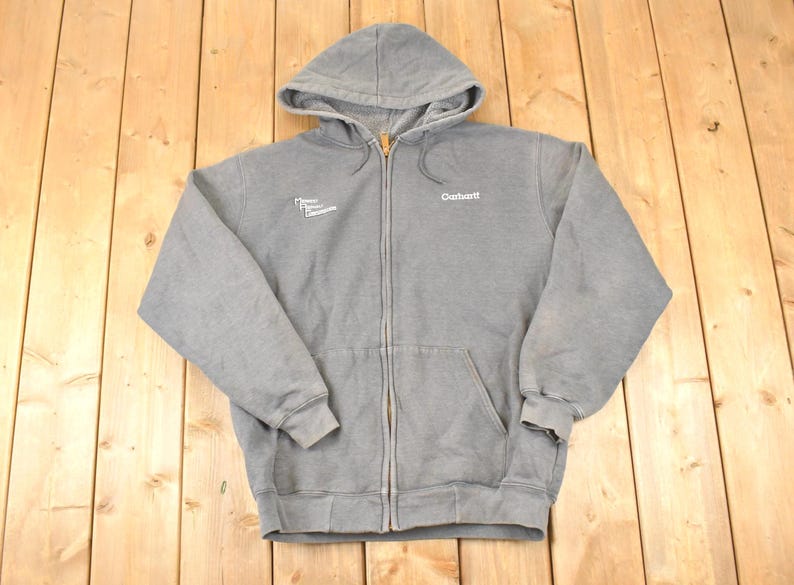 Puede incluir: Una sudadera gris con cremallera, capucha y bolsillo delantero. El logotipo de Carhartt es visible en el pecho derecho. La sudadera tiene un cord&oacute;n y pu&ntilde;os y dobladillo acanalados. El texto "Power Equipment Company" est&aacute; en el pecho izquierdo.