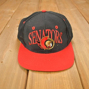 May include: Black and red baseball cap with the word "Senators" embroidered in red on the front. The cap features a gold and red logo of a winged Roman warrior.