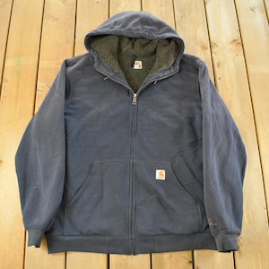 Puede incluir: Chaqueta con capucha azul marino con cremallera y forro polar. La chaqueta tiene un bolsillo delantero, una cremallera completa y un peque&ntilde;o parche con el logotipo. La chaqueta est&aacute; colocada sobre una superficie de madera.