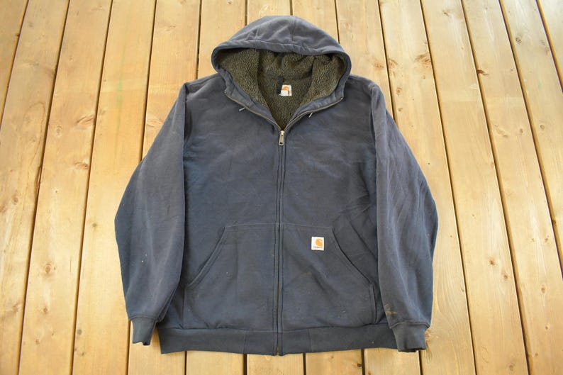 Puede incluir: Chaqueta con capucha azul marino con cremallera y forro polar. La chaqueta tiene un bolsillo delantero, una cremallera completa y un peque&ntilde;o parche con el logotipo. La chaqueta est&aacute; colocada sobre una superficie de madera.