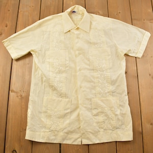 May include: A pale yellow short-sleeved shirt with white embroidery. The shirt has a button-up front and two pockets.