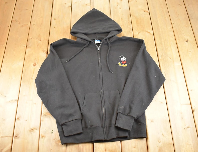 May include: A black zip-up hoodie with a hood and front pockets. The hoodie features a small embroidered image of Mickey Mouse on the left chest. The garment is made of a soft material and has a drawstring at the hood.