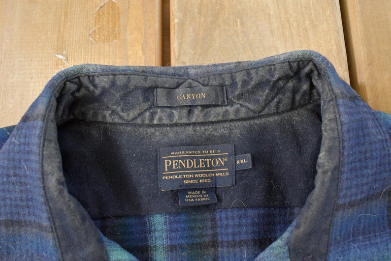 May include: Detail of a blue and green plaid shirt collar. The collar features a dark blue quilted lining and a small tag reading "CANYON". Below, a label states "PENDLETON WOOLEN MILLS" and "XXL".