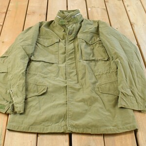 May include: An olive green military-style jacket with a button-up front and several pockets. The jacket features a high collar and long sleeves. The garment is made of a robust fabric and is displayed on a wooden surface.