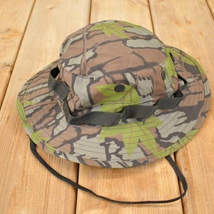 Può includere: Cappello da boscaiolo con motivo mimetico, con tesa larga e sottogola. Il cappello presenta un mix di colori marrone, verde e beige, creando un design mimetico boschivo. Il cappello è realizzato in tessuto resistente.