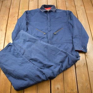 Puede incluir: Un traje de trabajo azul marino, compuesto por una chaqueta y un pantalón a juego, descansa sobre una superficie de madera. La chaqueta presenta un cuello marrón con forro rojo, dos cremalleras doradas y puños abotonados. El pantalón está doblado.