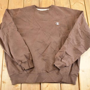 May include: A brown Champion sweatshirt with a crew neck and long sleeves. The sweatshirt features a small white Champion logo on the chest. The fabric appears to be a soft, worn material, and the garment is laid flat on a wooden surface.