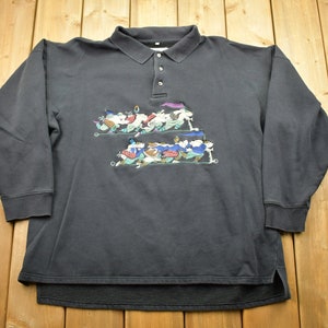 May include: A long-sleeved black polo shirt featuring a colourful cartoon graphic of a group of people riding on a sled. The graphic is centred on the shirt and features a variety of colours, including blue, green, red, and yellow.