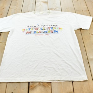 Op de afbeelding: Een wit T-shirt met korte mouwen en een kleurrijke afbeelding op de borst met de tekst Grand Opening TEEN CENTER MILLENNIUM JUNE 12, 1999. Het ontwerp bevat kleine illustraties van rode, gele, blauwe en groene ballonnen rondom de tekst.
