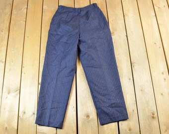 May include: Navy blue pinstripe trousers. The pants are made of a lightweight fabric and feature a classic pinstripe pattern. The trousers have a straight leg cut and a high waist. The pants are laid flat on a wooden surface.