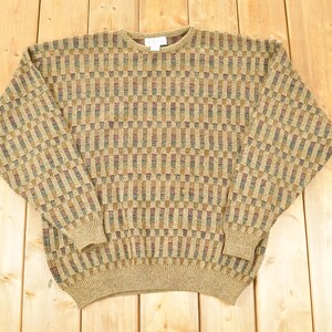 Könnte beinhalten: Ein beigefarbener Pullover mit einem geometrischen Muster in Braun-, Grün- und Blautönen. Der Pullover hat einen Rundhalsausschnitt, lange Ärmel sowie gerippte Bündchen und Saum. Das Kleidungsstück ist aus weichem, strukturiertem Strick gefertigt.