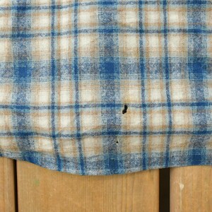 May include: Close-up of a blue, tan, and white plaid fabric. The pattern features a grid of intersecting lines, creating a classic check design. The fabric appears to be a woven material, possibly cotton or a similar textile. There are a few small holes in the fabric.