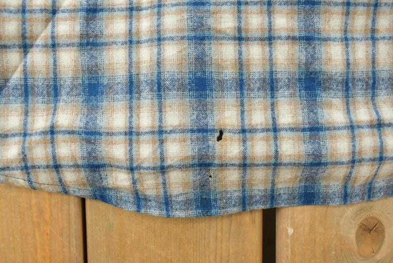 May include: Close-up of a blue, tan, and white plaid fabric. The pattern features a grid of intersecting lines, creating a classic check design. The fabric appears to be a woven material, possibly cotton or a similar textile. There are a few small holes in the fabric.
