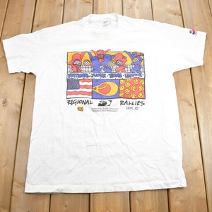 Puede incluir: Camiseta blanca con un gr&aacute;fico colorido de jugadores de tenis y el texto "National Junior Tennis League" y "Regional Rallies 1992".