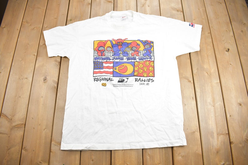 Puede incluir: Camiseta blanca con un gr&aacute;fico colorido de jugadores de tenis y el texto "National Junior Tennis League" y "Regional Rallies 1992".