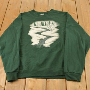 Puede incluir: Una sudadera de manga larga de color verde oscuro con un estampado gráfico central de un paisaje invernal. El diseño muestra un río sinuoso que fluye a través de un suelo cubierto de nieve con un bosque de pinos al fondo, en blanco y gris.