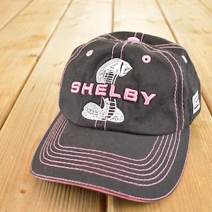 Op de afbeelding: Zwarte baseballcap met roze stiksels en een roze "SHELBY" logo met een cobra-ontwerp. De cap heeft een gebogen klep en een gestructureerde pasvorm.