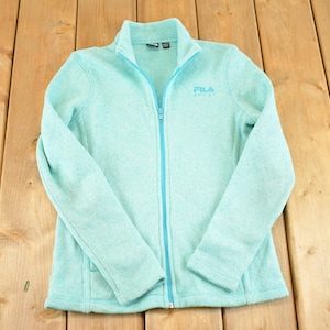 May include: A light blue Fila fleece jacket with a full zipper, long sleeves, and a stand-up collar. The Fila logo is embroidered on the chest. The jacket is laid flat on a wooden surface.