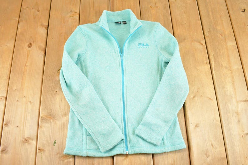 May include: A light blue Fila fleece jacket with a full zipper, long sleeves, and a stand-up collar. The Fila logo is embroidered on the chest. The jacket is laid flat on a wooden surface.