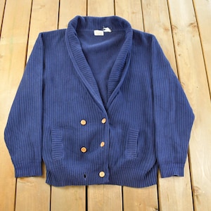 May include: A navy blue double-breasted cardigan sweater with a shawl collar and ribbed texture. The sweater features two front pockets and gold-colored buttons. A tag is visible near the collar. The garment is laid flat on a wooden surface.