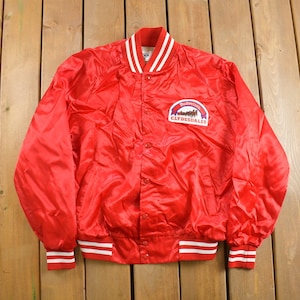 Puede incluir: Una chaqueta bomber de satén rojo con ribetes blancos y rojos alrededor del cuello, los puños y la cintura. La chaqueta presenta un parche con el logotipo de Budweiser Clydesdales en el pecho izquierdo.