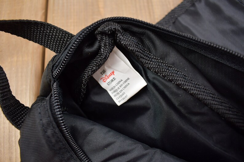 Op de afbeelding: Zwarte ritszak met een rood en wit label met de tekst "The Disney Store" en "Gemaakt in China" in het Engels, Frans en Chinees.