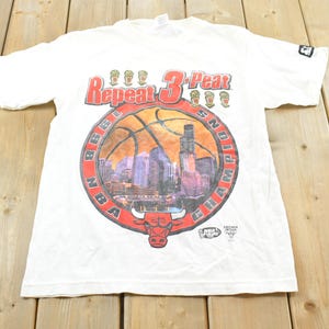 May include: White t-shirt featuring a basketball design with the text "Repeat 3-Peat" in red. The design includes a cityscape and a bull logo, with "NBA 1998 Champions" around the basketball. The shirt has short sleeves.