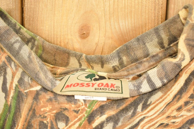 May include: Close-up of a camouflage pattern shirt collar. The shirt features a brown, green, and tan pattern. The tag reads "Mossy Oak Brand Camo". The shirt is made of a soft, comfortable material.