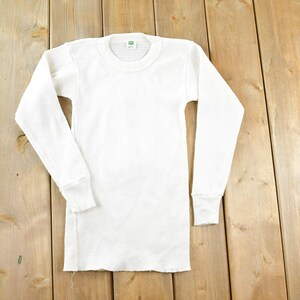 May include: A white long-sleeved thermal top with a crew neck and ribbed cuffs. The top is made of a textured fabric and is laid flat on a wooden surface.