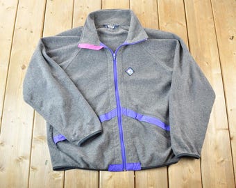 Puede incluir: Chaqueta de forro polar gris con cremallera completa, con detalles en morado y rosa. La chaqueta tiene un cuello alto y un pequeño emblema en el pecho. La cremallera y los ribetes son morados, y el cuello tiene detalles rosas.