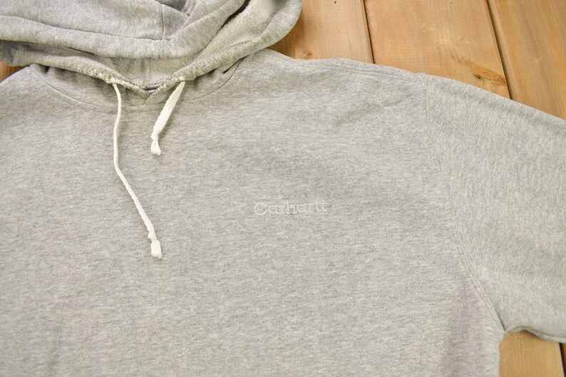 Puede incluir: Sudadera gris con capucha y un logotipo blanco de Carhartt bordado en la parte delantera.