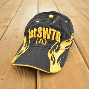 Op de afbeelding: Zwarte baseballcap met gele geborduurde tekst "1stSWTG (A)" en vlamontwerpen. De cap heeft een gebogen klep en een gele knop. Een modieus accessoire voor casual wear.