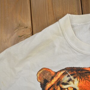 K&ouml;nnte beinhalten: Wei&szlig;er Pullover mit einem Tiger-Grafikdruck. Der Pullover hat einen Rundhalsausschnitt und ein Jerzees-Markenetikett.