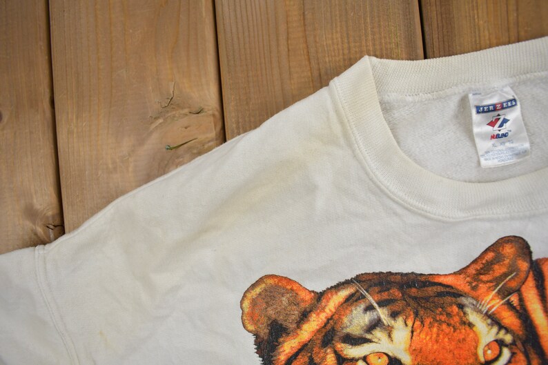 K&ouml;nnte beinhalten: Wei&szlig;er Pullover mit einem Tiger-Grafikdruck. Der Pullover hat einen Rundhalsausschnitt und ein Jerzees-Markenetikett.