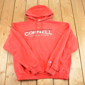 Puede incluir: Sudadera con capucha roja con el texto blanco "CORNELL THE HOTEL SCHOOL". El logo de Champion es visible en la manga izquierda. La sudadera es de material suave y está sobre una superficie de madera.