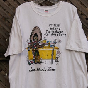 May include: White t-shirt with a cartoon illustration of a man wearing a sombrero and holding a gun. The text on the shirt reads "I'm Quiet I'm Happy I'm Handsome I don't give a Chit!!" and "San Antonio, Texas".