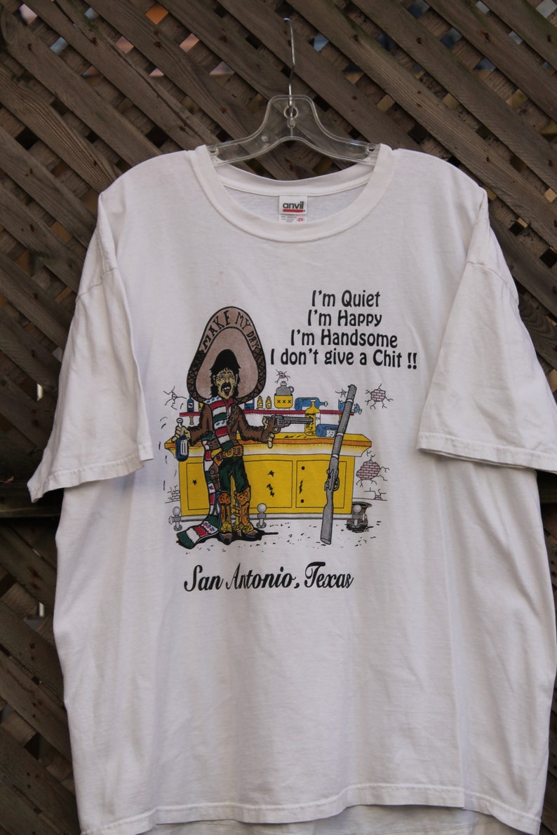 May include: White t-shirt with a cartoon illustration of a man wearing a sombrero and holding a gun. The text on the shirt reads "I'm Quiet I'm Happy I'm Handsome I don't give a Chit!!" and "San Antonio, Texas".