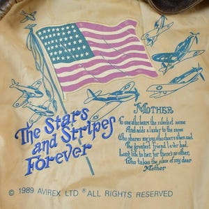 May include: Close-up of the inside lining of a vintage bomber jacket. The lining is cream-colored with a design featuring an American flag, airplanes, and the text "The Stars and Stripes Forever." The text "Mother" is also present.