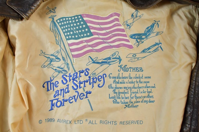 May include: Close-up of the inside lining of a vintage bomber jacket. The lining is cream-colored with a design featuring an American flag, airplanes, and the text "The Stars and Stripes Forever." The text "Mother" is also present.