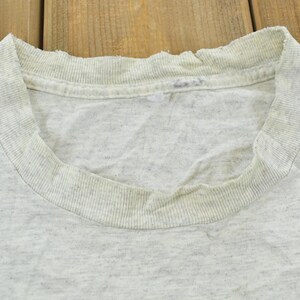 Op de afbeelding: Een lichtgrijs gem&ecirc;leerd T-shirt met ronde hals en een geribbelde kraag met zichtbare gebruikssporen, waaronder rafeling langs de rand van de halslijn en lichte verkleuring rond de binnenste kraagband, uitgevoerd in een klassiek model.