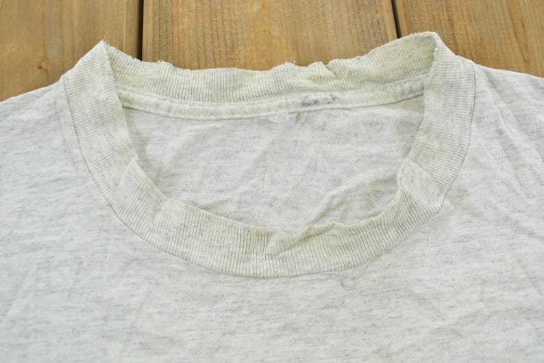 Op de afbeelding: Een lichtgrijs gem&ecirc;leerd T-shirt met ronde hals en een geribbelde kraag met zichtbare gebruikssporen, waaronder rafeling langs de rand van de halslijn en lichte verkleuring rond de binnenste kraagband, uitgevoerd in een klassiek model.
