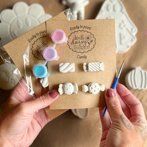 May include: A craft kit featuring four small white plaster candy shapes attached to a brown card labeled Ready to paint do da daisy Candy. The kit includes a strip of three small paint pots in pink, purple, and blue, and a thin blue paintbrush.