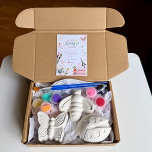 Op de afbeelding: Open kartonnen doos met een doe-het-zelf-schilderset. De set bevat een vlinder, bij en lieveheersbeestje beeldjes, verfpotjes in verschillende kleuren en penselen. Een kaart met de tekst "Thank You!" is ook zichtbaar.