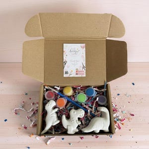Könnte beinhalten: Geöffnete Pappschachtel mit drei weißen Dinosaurierfiguren, Farbtöpfen in verschiedenen Farben, einem Pinsel und bunten Konfetti. Eine kleine Karte mit Text ist in der Schachtel sichtbar. Die Schachtel steht auf einer hellen Holzoberfläche.