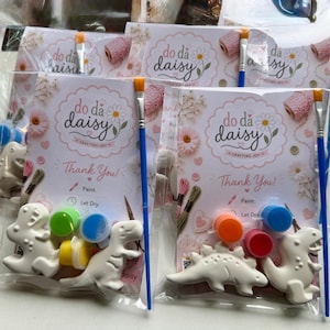 May include: Packaged DIY dinosaur painting kits. Each kit includes white ceramic dinosaur figures, paint pots in various colours, paintbrushes, and a card with the text "do da daisy" and "Thank You!".