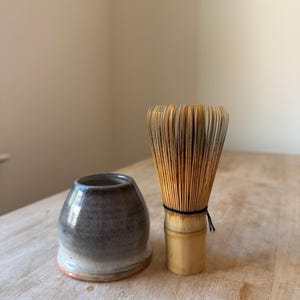 May include: A bamboo matcha whisk with thin, light brown tines and a cylindrical handle, positioned next to a small, rounded ceramic whisk holder featuring a gradient of dark gray and white glaze with a visible terracotta-colored base rim.